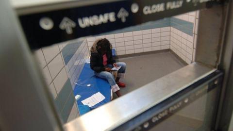 A police cell viewed through a hatch with a person sitting on a bed within. 