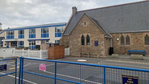 Castel Primary School Guernsey. It two distinctly different buildings side by side, separated by a fenced playground area. n the right is an older stone building constructed from rough, brownish masonry. It has a steep, slate-grey roof and several narrow, arched windows. To the left is a much more modern two‑storey structure with a cream-colored exterior and bright blue window frames.