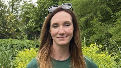 A woman in a green top with long straight hair and sunglasses on her head, smiles warmly while standing in front of green foliage and trees.