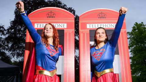 Two women wearing Superman costumes. Each raises one arm in the air in a superhero pose. They stand in front of red telephone boxes.