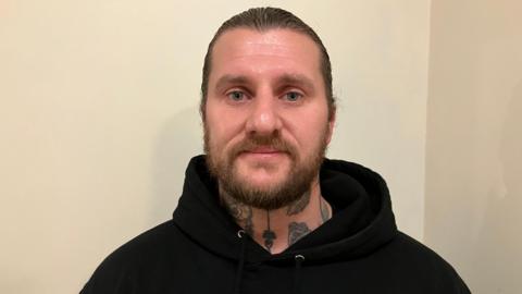 A man with a beard and hair pulled back is wearing a black hoodie, and has tattoos on his neck.