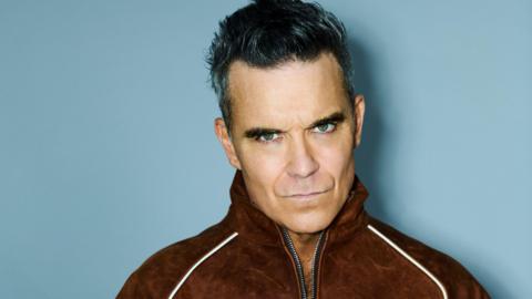 Robbie Williams looks at the camera with a serious expression, with greying black hair and wearing a brown tracksuit top