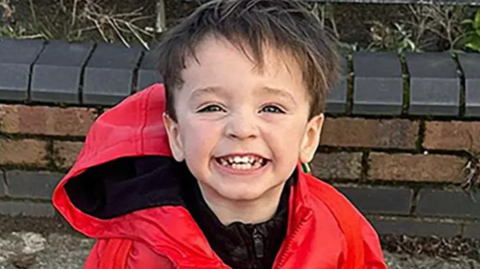 Jude Gerrard smiling widely at the camera. He has short brown hair and is wearing a bright red coat with the hood down