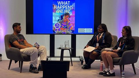 Two school pupils sit carrying out an interview on a stage, with Nathanael Lessore sitting across from them holding a book. A table with water on it is in-between, and a poster advertising the man's book - What Happens Online is behind them.
