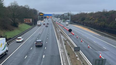 A view of the M1 looking northbound near junction 13 for Brogborough in Bedfordshire. All lanes are open northbound but the outside lane of the southbound stretch is closed by a series of cones and signage. It is a cloudy, wet day.