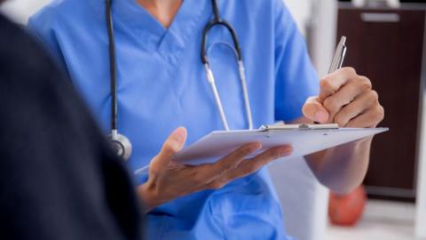 Nurse in blue scrubs wearing a black stethoscope, her face is cropped out of the photo. She is holding a clipboard writing with a silver pen. The background is blurred around her. 