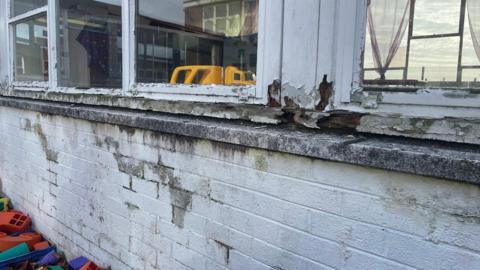 A discoloured white brick wall and window. The window frames are white but the paint is crumbling off. Some colourful toys can be seen in the bottom left of the picture on the ground laying against the wall. Another yellow toy can be seen through the window as can a reflection of another part of the building.  