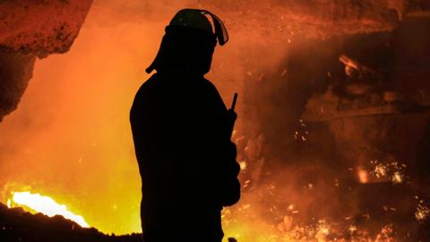 Worker at steel plant stands silhouetted against burning fires 