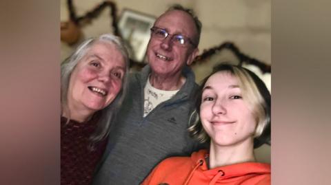 A selfie photo shows three people standing close together. There is a woman with grey hair and a purple jumper on the left, a man with grey hair and a grey sweater in the middle, and a person with short brown hair that has bleached blonde streaks on the right.