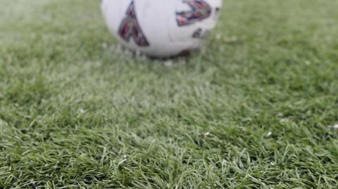 A close-up shot of a football resting on an artificial grass pitch. The pitch is green and focused on it with the white football out of focus in the distance. 