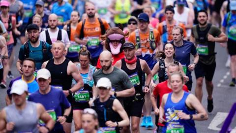 A large group of runners moving along a city street during a race. Participants are wearing colourful athletic gear, race bibs with numbers, and hydration packs. Among the crowd, one runner stands out wearing a full animal costume resembling a brown bull with horns and a pink snout.