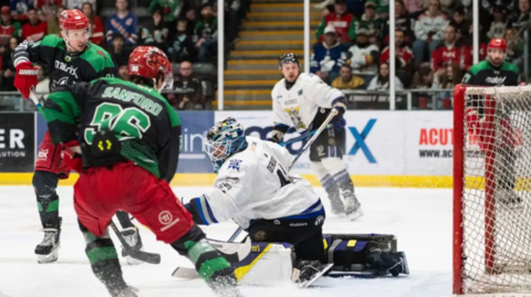 Storm's goalkeeper, wearing white and a black helmet, and an attacker in red trousers and a black and green top, fight for the puck on the ice in front of the net
