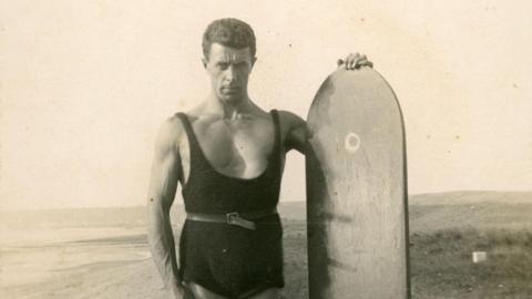 An old photo of Nigel Oxenden wearing a swim suit standing holding a surfboard. In the background is a beach. 