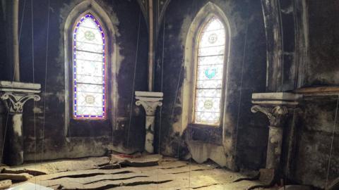 Inside the monastery in Paignton. There are two church windows at the back and the building is dark. The floor is damaged.