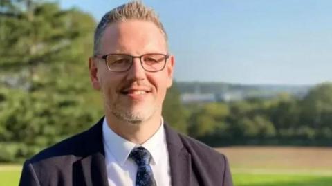 A man with spiky short hair and glasses wearing a suit with a black and blue floral tie standing outside with trees in the background