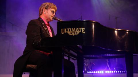 Ultimate Elton, an Elton John tribute act, with a man sitting at a piano singing and dressed like Elton John. He is wearing a glittery jacket, sunglasses and an earring and the stage is lit up in purple.