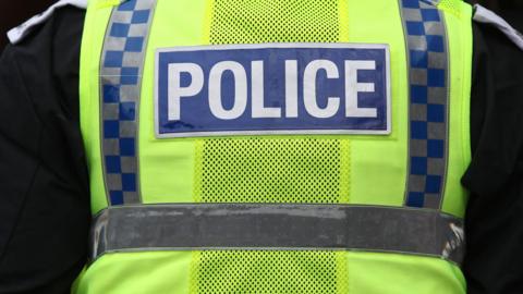 A stock image of a police officer seen from the back. They are wearing a yellow high visibility vest with the word 'police', over a black jacket.