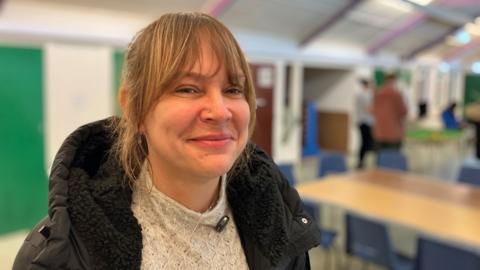 Clare Lincoln, a youth manager, who is standing inside the room of a youth club. She has her hair pulled back off her face and a full fringe and is looking directly at the camera and smiling. She is wearing a beige jumper with a black coat on.