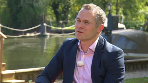 Tim Mcmahon has short hair and is wearing a pink shirt and a blue jacket. He is sat in front of the River Thames, which is flowing by in the sun. 