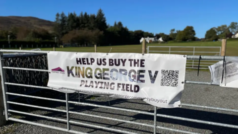 A white fabric sign attached to a metal farm-style gate. The sign has a message that reads: "Help us buy the King George V playing field.