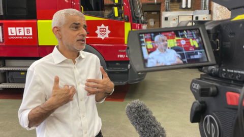 Khan being interviewed inside a fire station, speaking in front of a red London Fire Brigade truck while a camera and microphone record the scene.