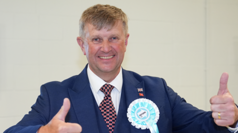 Peter Harris, wearing a blue suit with a white shirt and a patterned tie, smiles at the camera as he gives the thumbs up.