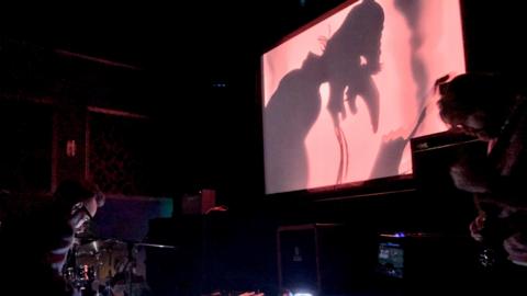 A man hunched over a drum kit and another man playing a string instument play music in a dark room. A screen is above them showing an animated film, with the film showing a cartoon of a man with a long nose and beard looking down.