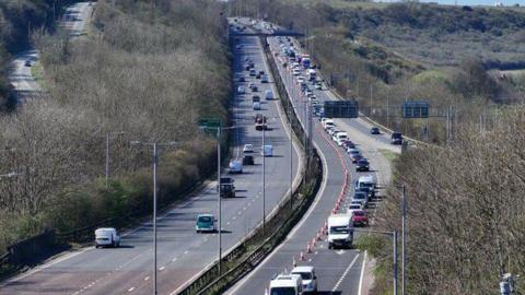 A two carriage A road. The carriageway on the left has one lane closed by cones and congested traffic. 