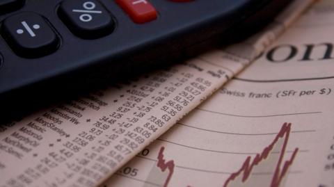 A calculator lies on top of the financial pages of a newspaper which shows a chart with a rising and falling red line and columns of share prices.