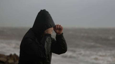A man wearing a black hooded raincoat trying to protect his face from the rain and tide - pictured in the background.