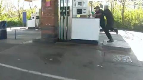 cctv footage of a petrol station forecourt. In the top right corner a police officer can be seen tackling a man doused in petrol holding a lighter