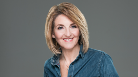 Kaye Adams smiling straight at the camera. She is wearing a blue shirt and is standing against a grey background.