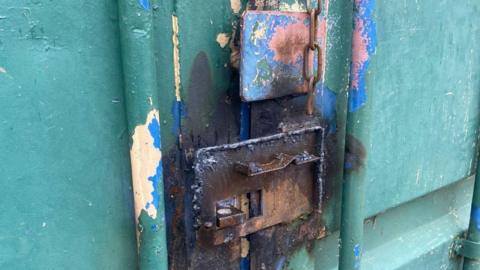 A close-up of a lock on a green container showing it burnt, damaged and rusty, with the padlock missing.