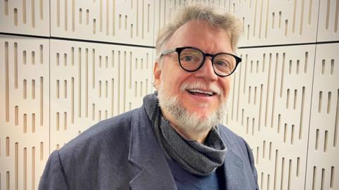 Guillermo Del Toro, wearing a grey jacket, scarf, and glasses