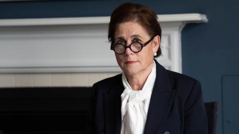 Baroness Lampard inside Arundel House in London, where the Lampard Inquiry is sitting until the end of 2026. She is wearing dark brown glasses which have rounded frames, pearl earrings and has short brown hair. She has a navy jacket on and a white blouse. She is sitting in front of a fireplace with a white hearth.