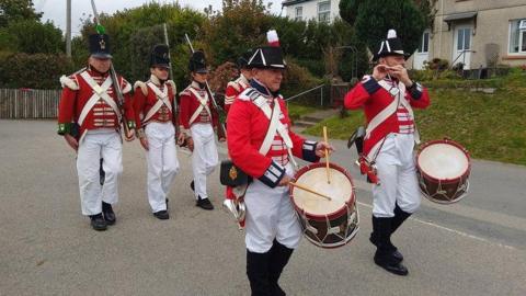 Members of the Plymouth Maritime Corps of Drums and members of the 32nd Cornwall Regiment of Foot, all in 1805 uniform, walk down the road.