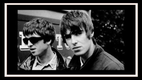 Black and white picture of Noel and Liam Gallagher.  Noel is wearing black sunglasses and looking away and Liam is looking down the lens of the camera.