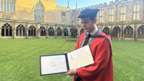 Matt Baker wearing a red graduation cloak, on top of a grey blazer with a maroon tie, holding up a two sided folder which holds his certificate and documents. 
