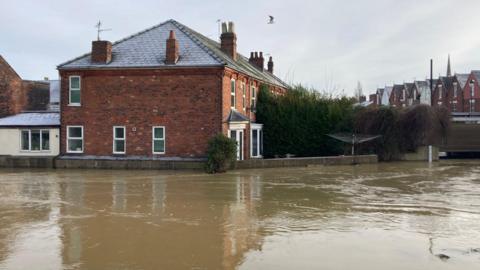 A residential street will with brown flood water in front of a row of terraced red-brick houses.