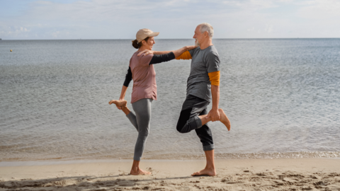 A woman and man reach an arm to the other's shoulder for balance, whilst standing on one foot on a beach.