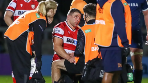 A Gloucester Rugby player, wearing a red and white striped kit, talks to pitch side medical professionals, wearing orange tabards, on the pitch under the lights following an injury