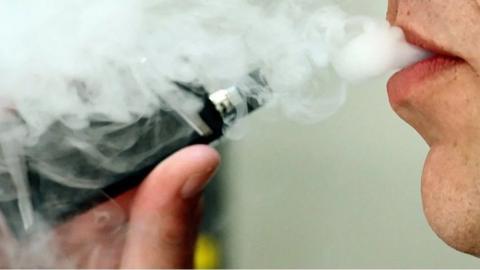 Close up of a person holding a vape near to their mouth and exhaling a large amount of mist 