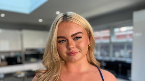 The young women has long, blonde hair falling over her right shoulder. She is wearing a blue dress with thin straps and gold hoop earrings. She has tanned skin and blue eyes and is looking straight into the camera with a closed-mouth smile.