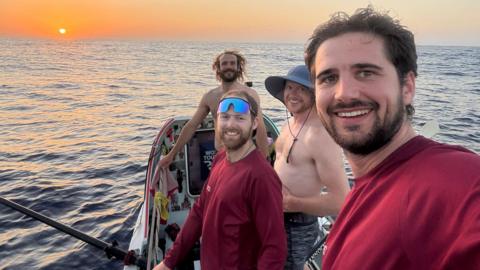 Four men smile at the camera on a rowing boat in open sea.