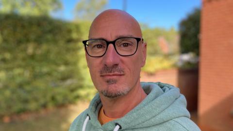 A man looks toward the camera he is wearing black rimmed glasses and has a grey goatee beard, he is wearing a light green hoodie behind him is a hedge and a red brick building.