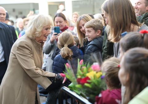 Queen Camilla wears a brown jacket and shakes hands with a well-wisher with people watching her behind a fence