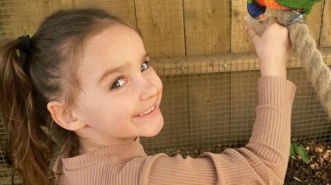 A girl with brown hair tied back in a ponytail smiles at the camera as she feeds a parrot.