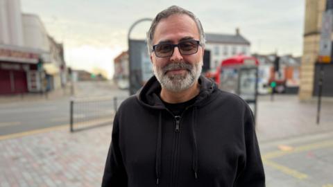 A man wearing a black hoodie and sunglasses stands in a street.