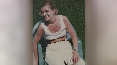 A picture of Joanne Tulip. She is sat on a green plastic garden chair in front of a hedge. She has blond hair in a ponytail and is wearing a cream crop top, cream trousers, golden earrings and bracelet and a watch. She is smiling at the camera.