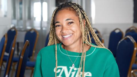 A woman with long braided hair smiling at the camera. She is wearing a green jumper with a Nike logo.